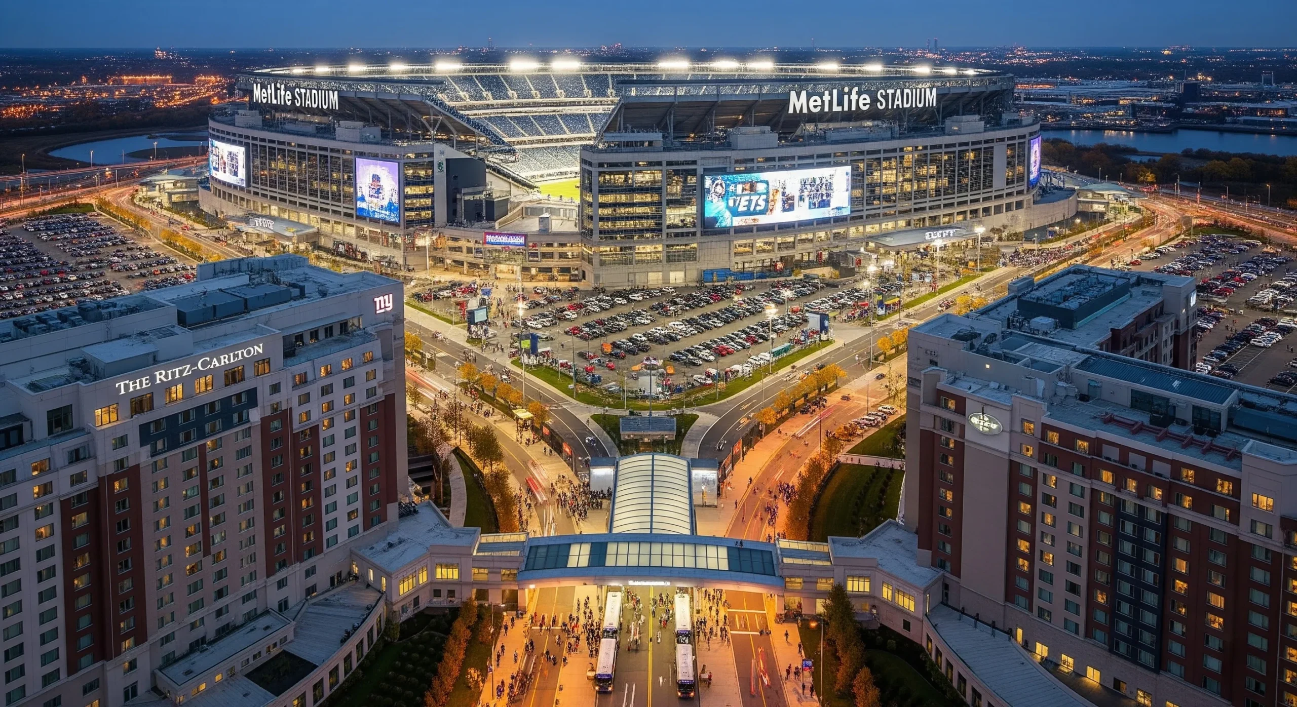 Aerial view of MetLife Stadium in East Rutherford with surrounding luxury and budget hotels, showing the massive NFL venue, parking areas, and New York City skyline in the distance